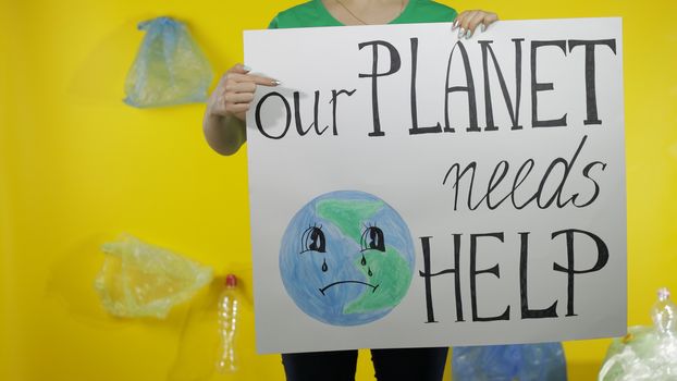 Unrecognizable woman volunteer in t-shirt with recycle logo holding protesting message poster Our Planet Needs Help. Background with cellophane bags, bottles. Environment trash plastic pollution