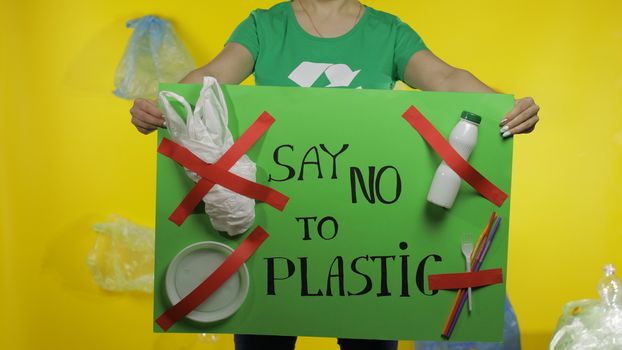 Unrecognizable woman activist in t-shirt with recycle logo holding protesting message poster Say No To Plastic. Background with cellophane bags, bottles. Environment trash plastic pollution