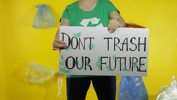 Unrecognizable woman volunteer in t-shirt with recycle logo holding protesting message poster Don't Trash Our Future. Background with cellophane bags, bottles. Environment trash plastic pollution