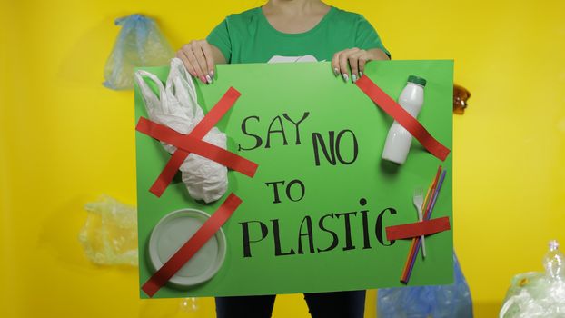 Unrecognizable woman volunteer in t-shirt with recycle logo holding protesting message poster Say No To Plastic. Background with cellophane bags, bottles. Environment trash plastic pollution