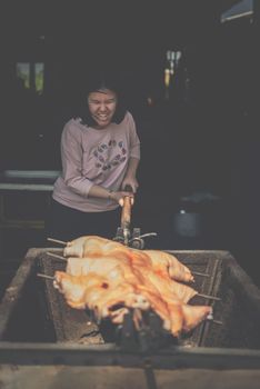 Asian woman chef cooking Barbecued Suckling Pig by roasting pork on charcoal for sale at Thai street food market or restaurant in Thailand