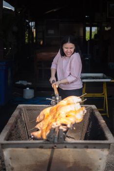 Asian woman chef cooking Barbecued Suckling Pig by roasting pork on charcoal for sale at Thai street food market or restaurant in Thailand