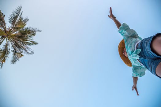 Low angle view of sexy woman short jeans midair by jumping, crossing step over camera shot below at beach with coconut and sky overhead in concept travel, active lifestyle, overcome obstacles in life