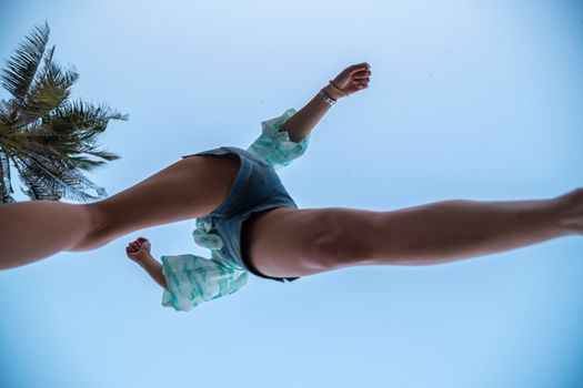 Low angle view of sexy woman short jeans midair by jumping, crossing step over camera shot below at beach with coconut and sky overhead in concept travel, active lifestyle, overcome obstacles in life