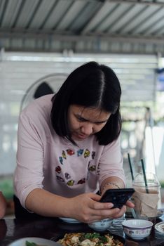 Asian woman foodstagramming use smartphone photographing a food on table in restaurant before eat for post and share on internet social media with fun and happy