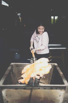 Asian woman chef cooking Barbecued Suckling Pig by roasting pork on charcoal for sale at Thai street food market or restaurant in Thailand