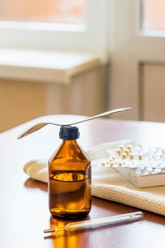 A cough syrup bottle with a spoon, a thermometer and some pills blister on the table close to the window