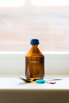 A cough syrup bottle with a spoon and some pills close to the window