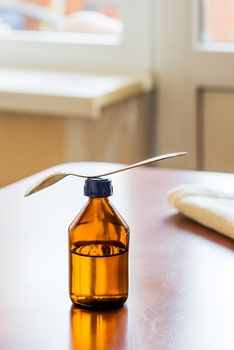 A cough syrup bottle with a spoon on the table close to the window