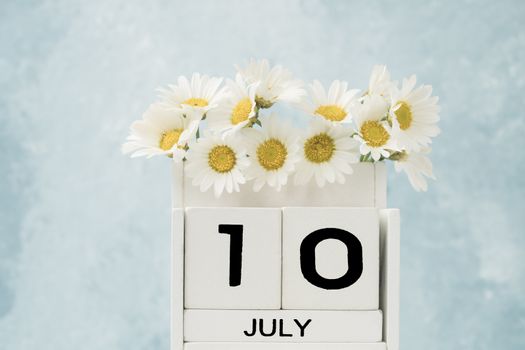 White cube calendar for july decorated with daisy flowers over blue background with copy space