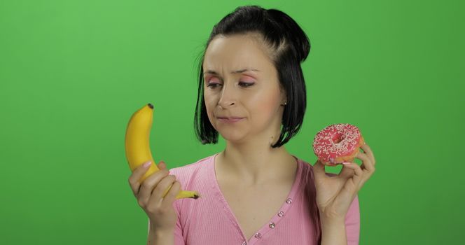 Starting healthy eating. Say no to junk food. Beautiful sad young girl on a chroma key background holds donut in one hand and banana on other. Cute woman choice donut or banana to eat