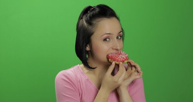 Happy beautiful young girl on a chroma key background with great pleasure eats a donut. Cute woman in a pink shirt posing with donut