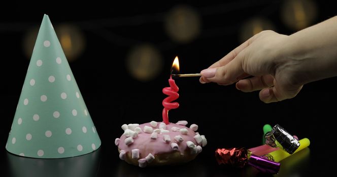 Party. Pink frosted donut with sprinkles and a red festive candle on it. Light the candle. Colored party hat. Black background. Celebrate an event, birthday. String of lights