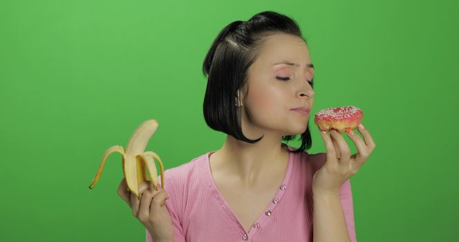 Starting healthy eating. Say no to junk food. Beautiful sad young girl on a chroma key background holds donut in one hand and banana on other. Cute woman choice donut or banana to eat