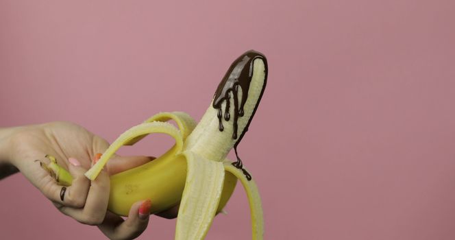 Banana with melted dark chocolate syrup. A peeled banana covered in chocolate cream. Pink background