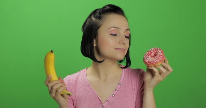 Starting healthy eating. Say no to junk food. Beautiful sad young girl on a chroma key background holds donut in one hand and banana on other. Cute woman choice donut or banana to eat