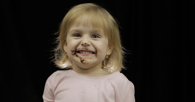 Beautiful, happy child eats melted dark chocolate syrup and whipped cream on black background. Teen blonde girl in pink shirt. Cute smiling. Dirty face
