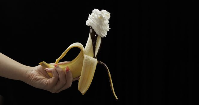 Woman hand holds banana with melted dark chocolate syrup and whipped cream on top of the fruit. A peeled banana covered in whipped cream. Black background