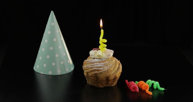Party. Cake and a yellow festive candle on it. Colored party hat. Black background. Celebrate an event, birthday