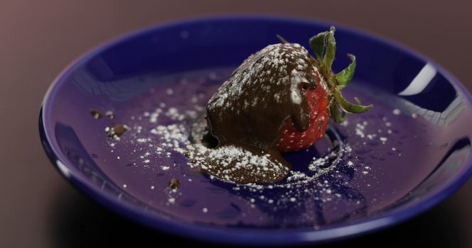 Ripe juicy strawberry in melted dark chocolate with icing sugar powder on a blue plate. Front view. Strawberry in chocolate on a dark background. Close up