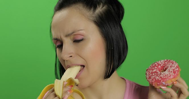 Starting healthy eating. Say no to junk food. Beautiful sad young girl on a chroma key background holds donut in one hand and banana on other. Cute woman choice donut or banana to eat