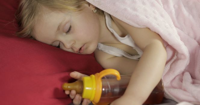 Cute baby girl sleeping on cozy bed at home with drinking bottle. Pretty, little girl sleeping in morning light. Close-up portrait of child naps. Concept of peaceful sweet dreams