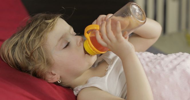 Cute baby girl sleeping on cozy bed at home and drinking juice from bottle. Pretty, little girl sleeping in morning light. Close-up portrait of child naps. Concept of peaceful sweet dreams