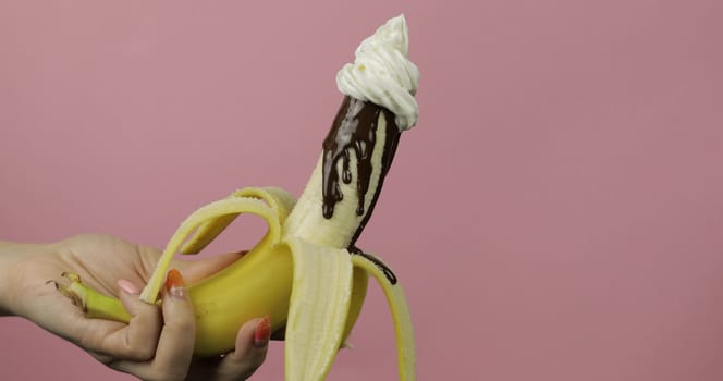 Banana with melted dark chocolate syrup and with whipped cream on top of the fruit. A peeled banana covered in chocolate cream. Pink background