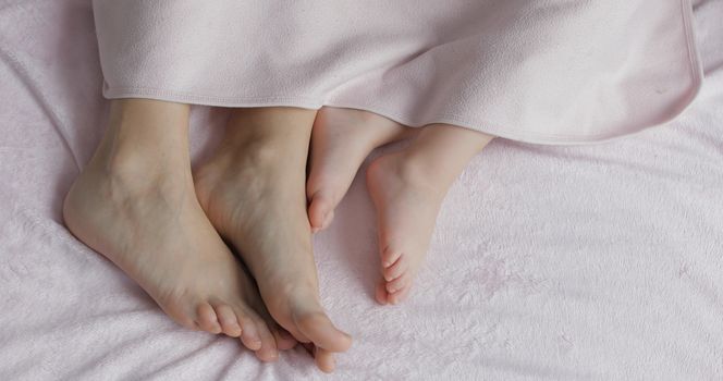 Two pairs of legs of the happy family in bed under the coverlet - mother and daughter. Sleeping of the baby and his mom on the bed in the morning. Happy family peaceful sweet dreams concept