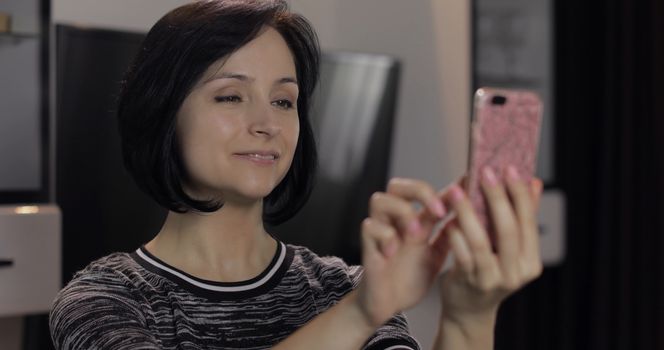 Portrait of pretty caucasian young brunette woman having chat on social media using smartphone enjoying chatting to friend. At home