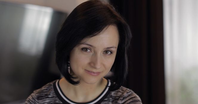 Portrait of pretty caucasian young brunette woman with brown eyes looking on the camera and smiling at home. Indoor. Smiling girl with cute face