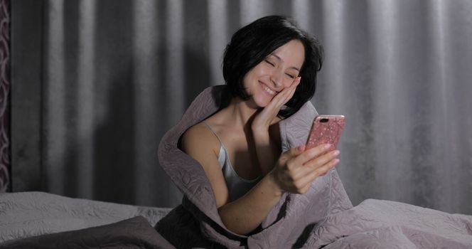 Pretty young brunette woman sitting on the bed under a blanket and having chat on social media using smartphone enjoying chatting to friend. Indoors. Blue nightgown. Online communication