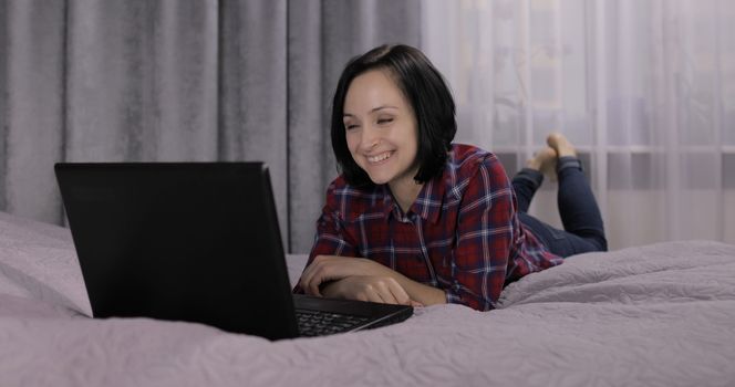 Pretty young brunette woman lying on the bed and having video chat using webcam on laptop computer enjoying chatting to friend. Indoors. Red shirt and jeans