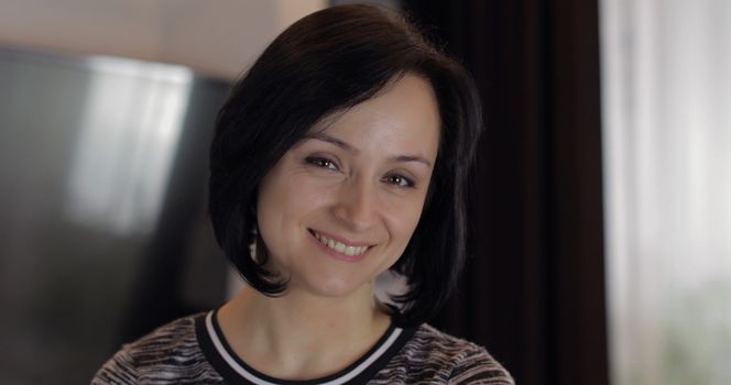 Portrait of pretty caucasian young brunette woman with brown eyes looking on the camera and smiling at home. Indoor. Smiling girl with cute face