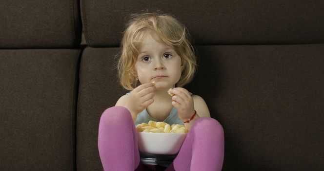 Beautiful little blonde girl sitting on a sofa at home and eating corn puffs. Cute child taste puffcorns. Inside. Childrens meal