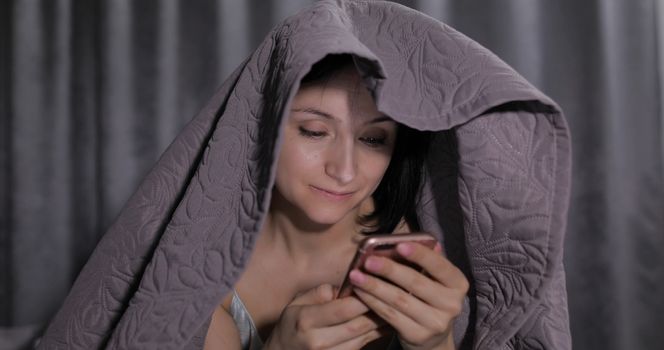 Pretty young brunette woman sitting on the bed under a blanket and having chat on social media using smartphone enjoying chatting to friend. Indoors. Blue nightgown. Online communication