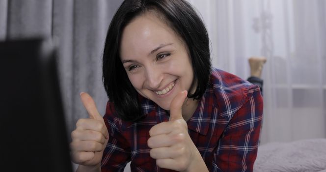 Pretty young brunette woman lying on the bed and having video chat using webcam on laptop computer enjoying chatting to friend. Indoors. Thumbs up. Red shirt and jeans