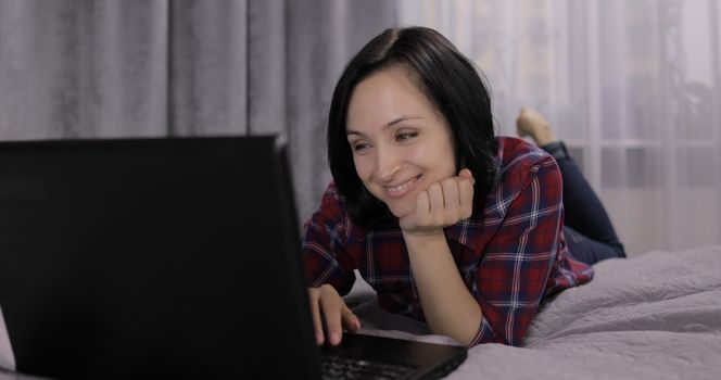 Pretty young brunette woman lying on the bed and watching something on the laptop computer. Chatting and working. Indoors. Red shirt and jeans