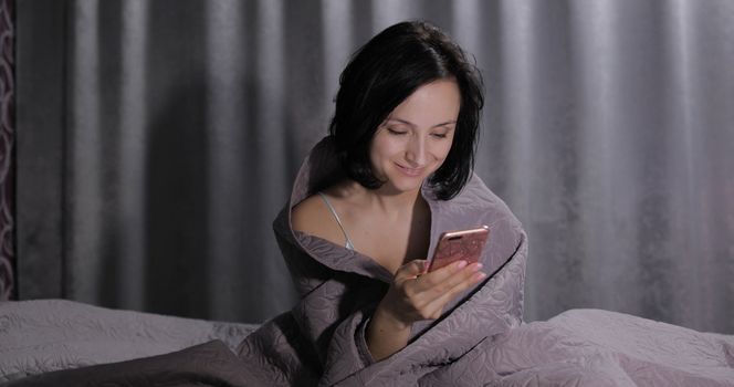 Pretty young brunette woman sitting on the bed under a blanket and having chat on social media using smartphone enjoying chatting to friend. Indoors. Blue nightgown. Online communication