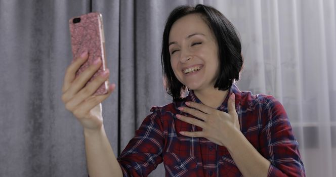 Pretty young brunette woman sit on the bed and having video chat using smartphone enjoying chatting to friend. Indoors. Red shirt and jeans. Online communication