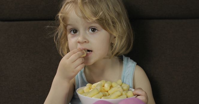 Beautiful little blonde girl sitting on a sofa at home and eating corn puffs. Cute child taste puffcorns. Inside. Childrens meal