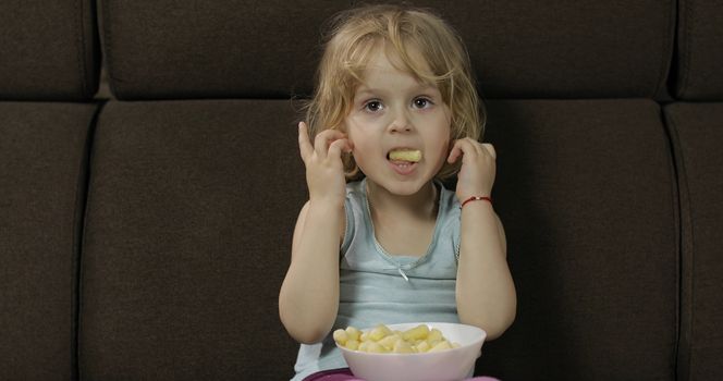 Beautiful little blonde girl sitting on a sofa at home and eating corn puffs. Cute child taste puffcorns. Inside. Childrens meal