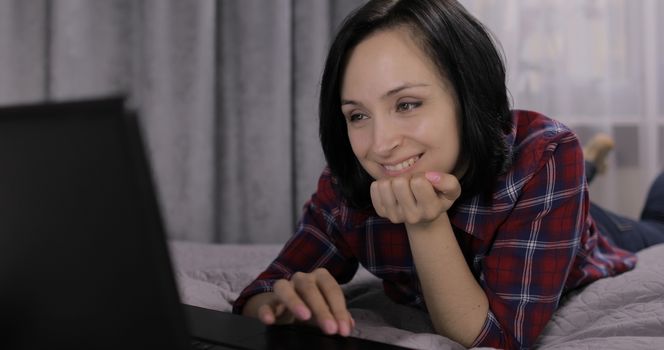 Pretty young brunette woman lying on the bed and watching something on the laptop computer. Chatting and working. Indoors. Red shirt and jeans