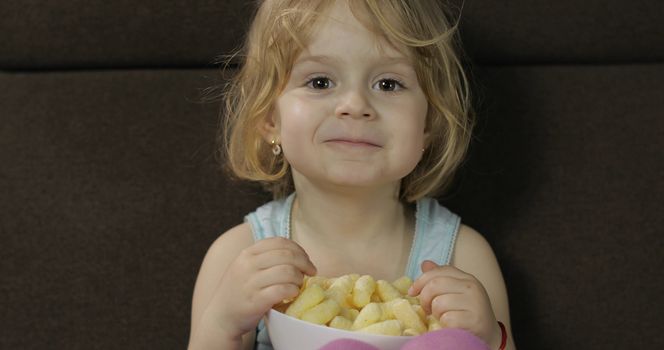 Beautiful little blonde girl sitting on a sofa at home and eating corn puffs. Cute child taste puffcorns. Inside. Childrens meal