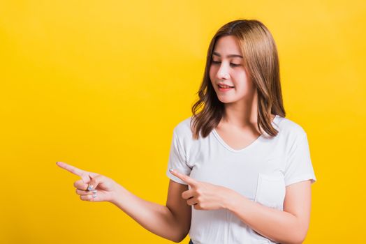 Asian Thai happy portrait beautiful cute young woman standing wear white t-shirt pointing finger away side looking to away side, studio shot isolated on yellow background with copy space