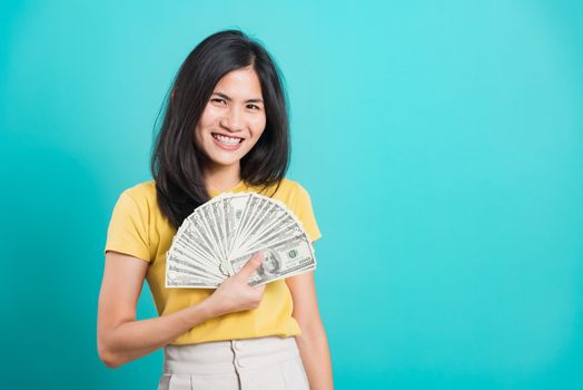 Asian happy portrait beautiful young woman standing wear t-shirt smiling holding money fan banknotes 100 dollar bills and looking to camera isolated on blue background with copy space for text