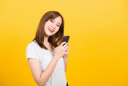 Asian happy portrait beautiful cute young woman teen smiling standing wear t-shirt using smart mobile phone looking to camera isolated, studio shot on yellow background with copy space