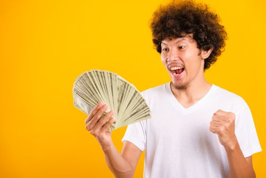 Asian handsome man with curly hair holding fans of money dollar bills isolate on yellow background with copy space for text
