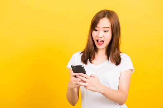 Asian happy portrait beautiful cute young woman teen smiling standing wear t-shirt her surprised excited with smart mobile phone looking the phone isolated, studio shot yellow background with copy space