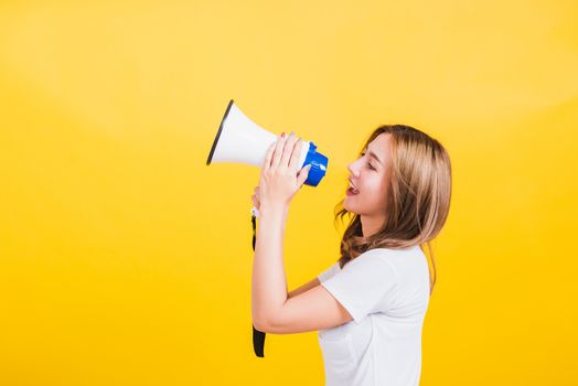 Asian Thai happy portrait beautiful cute young woman stand to make announcement message shouting screaming in megaphone looking to side away, studio shot isolated on yellow background with copy space